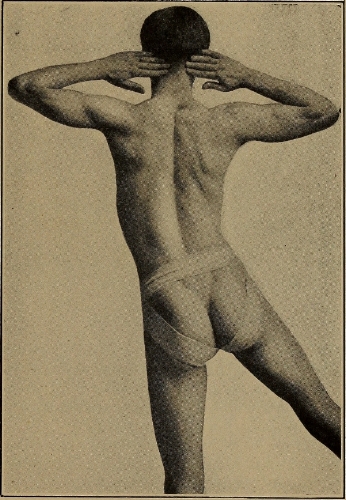 vintage photograph of the back of a man with his hands behind his head and lifting one leg off the ground to the side, like he is doing exercises. He is naked except for some kind of loincloth.