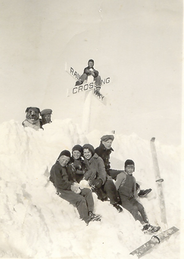 Vintage image of a group of youth sitting in a large snowbank. At the top of the snowbank, a 'RAILWAY CROSSING' sign sticks out of the snow. One boy sits atop the sign. There's also another boy and a dog half-submerged in the snow with their heads sticking out.